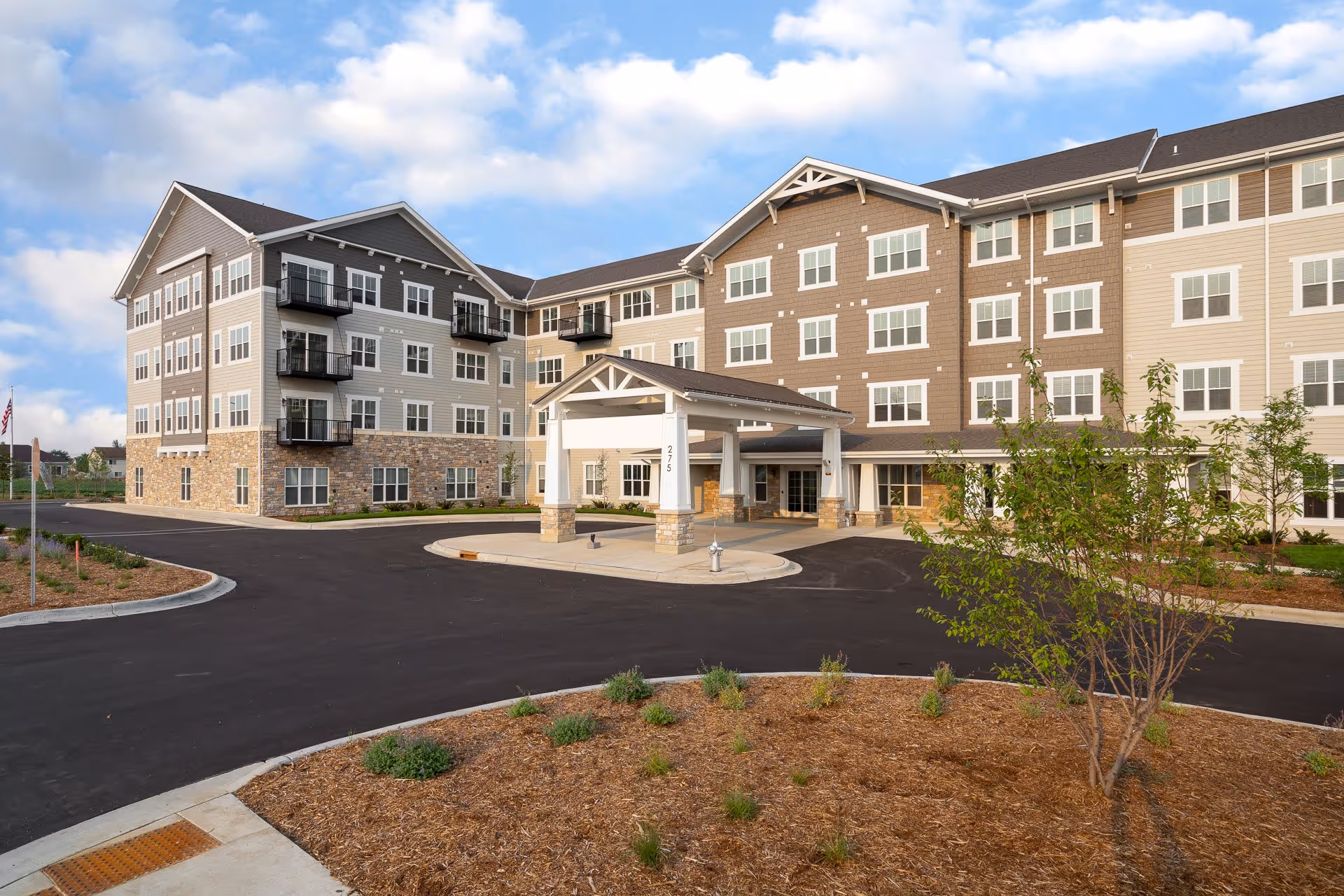 Exterior view of a modern senior living facility building with multiple floors, balconies, and a covered entrance driveway. The building has a combination of stone and siding on the facade, surrounded by landscaped areas with small plants and trees under a partly cloudy sky.