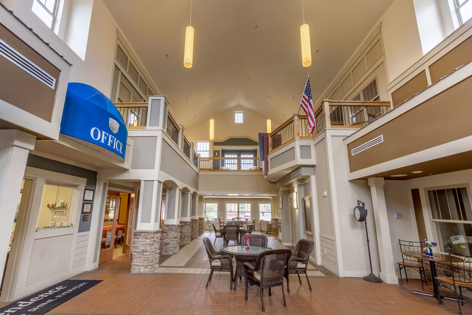 Spacious two-story senior living lobby with wicker seating, a blue office awning, and a vaulted ceiling with hanging lights.