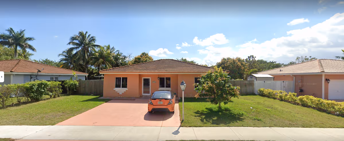 Single-story peach-colored house with a tiled roof, a driveway with an orange car parked on it, a small front yard with green grass and a tree, and neighboring houses on both sides under a clear blue sky with some clouds.