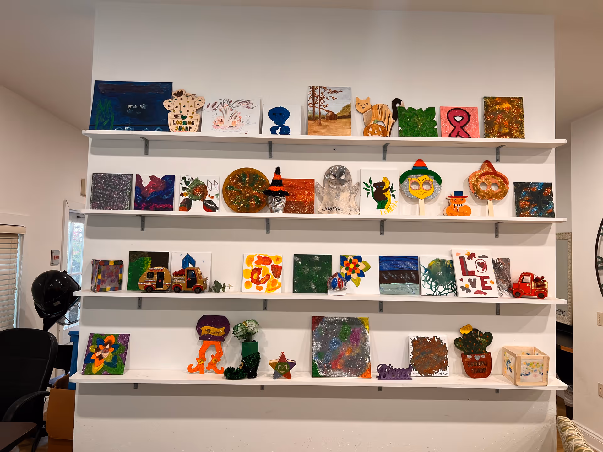 A white wall with four white shelves displaying various colorful handmade art pieces and crafts, including paintings, decorated wooden shapes, and small sculptures. The room has a chair and a black helmet on the left side, and a large clock partially visible on the right.