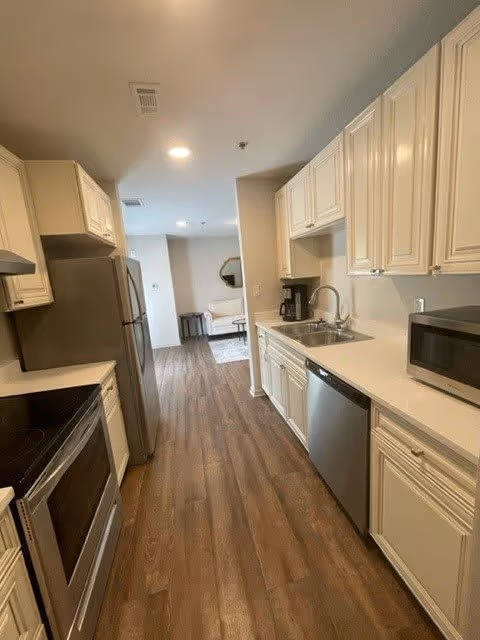 A modern kitchen with wooden flooring, white cabinets, stainless steel appliances including a refrigerator, dishwasher, microwave, and oven. The kitchen opens into a living area with a white sofa and a round mirror on the wall.