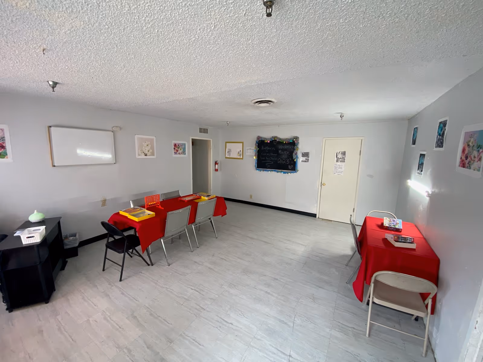 A simple activity room with two tables covered in red tablecloths. One table has six chairs around it and board games on top, while the other smaller table has two chairs and a puzzle. The walls are light gray with various floral and decorative pictures hanging. A blackboard on the far wall has a message about happiness and the date Wednesday, March 1, 2023. The floor is light-colored tile, and the ceiling has a textured finish with several light fixtures.