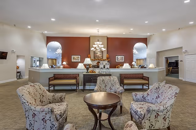 A spacious senior living facility common area with four floral-patterned armchairs arranged around a round wooden table. Behind the seating area is a half wall with two benches and a console table decorated with lamps and autumn-themed decor. The back wall is painted a warm red and features a chandelier and framed artwork. There are arched doorways on either side leading to other rooms.
