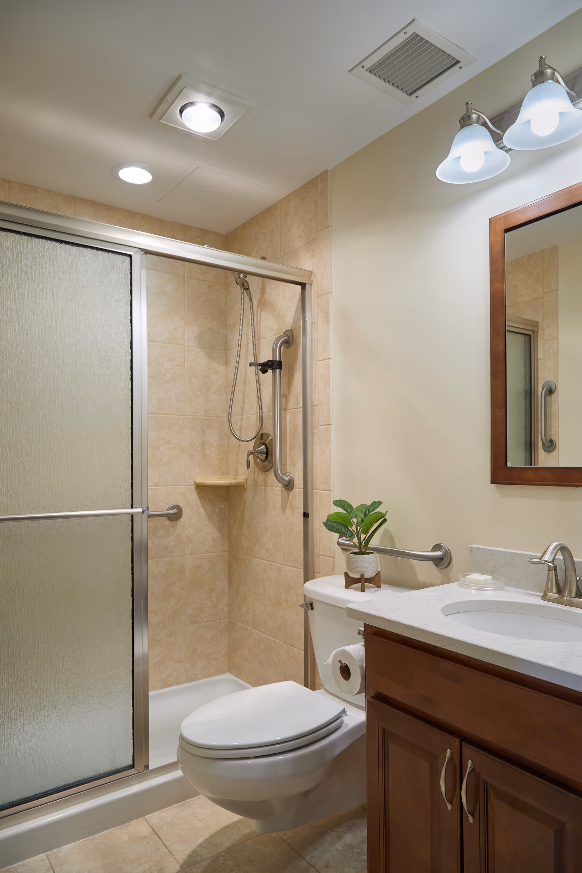 A clean and well-lit bathroom featuring a shower with sliding frosted glass doors and beige tiled walls. The shower has a handheld showerhead and a grab bar. In front of the shower is a white toilet with a small green potted plant on the tank. To the right is a wooden vanity with a white countertop, a sink, a faucet, and a mirror above it. The bathroom has light-colored walls and floor tiles.