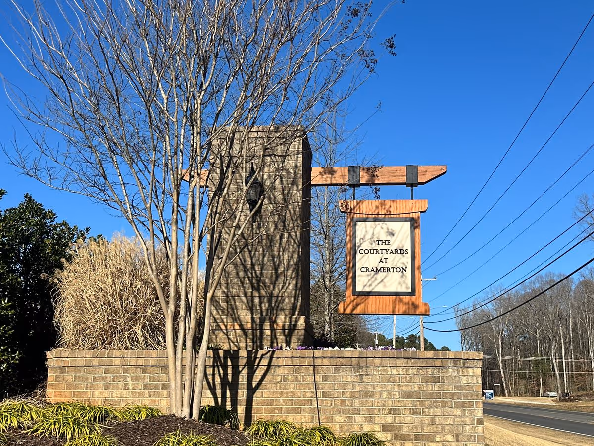 Brick entrance sign with a wooden hanging plaque reading 'The Courtyards at Cramerton' by a road under a clear blue sky.