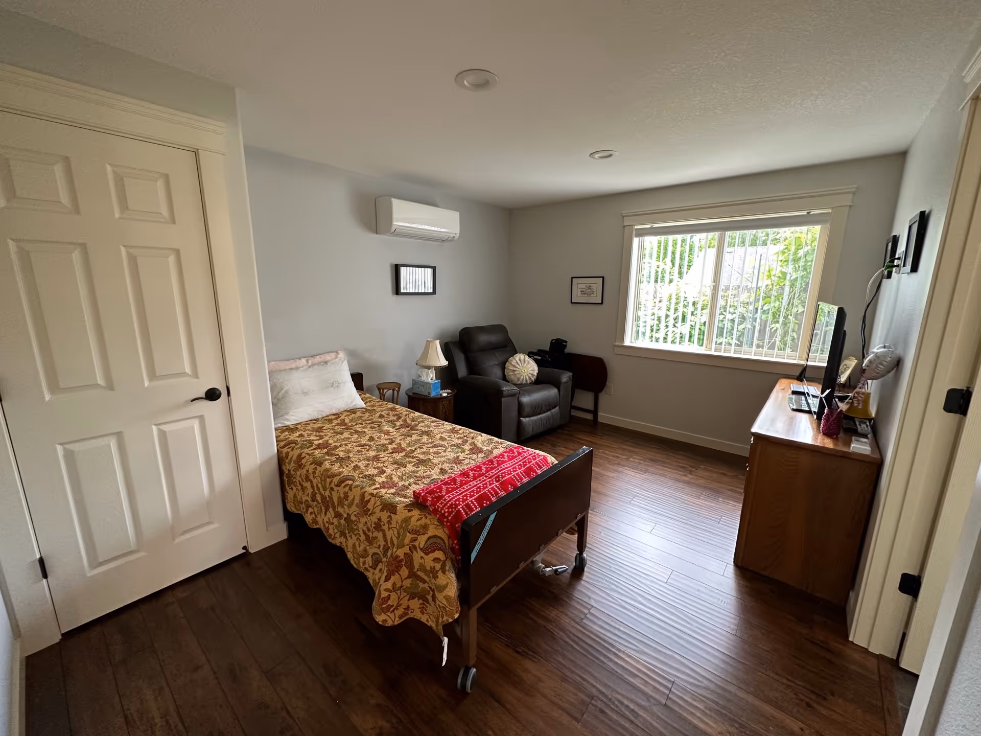 A cozy bedroom with a single bed covered in a floral patterned bedspread and a red blanket at the foot. Next to the bed is a small wooden nightstand with a lamp and a box of tissues. There is a dark recliner chair with a round pillow beside the nightstand. A large window with vertical blinds lets in natural light, and a wooden dresser with a TV on top is positioned against the opposite wall. The room has wood flooring and light-colored walls with minimal decorations.