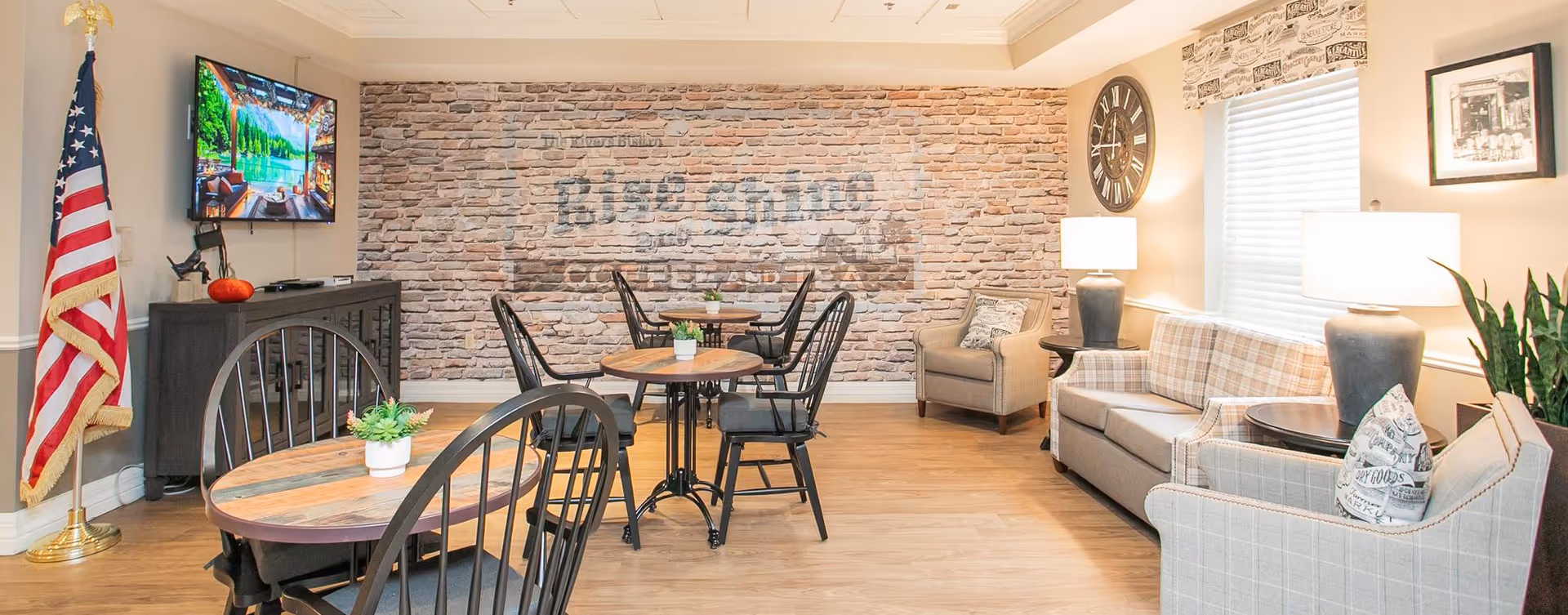 A cozy common area with wooden floors, featuring round wooden tables with black metal chairs, a plaid sofa, an armchair, and a large wall clock. A flat-screen TV is mounted on a cabinet against a brick accent wall with faded text. An American flag stands in the corner, and there are table lamps and potted plants adding to the warm ambiance.