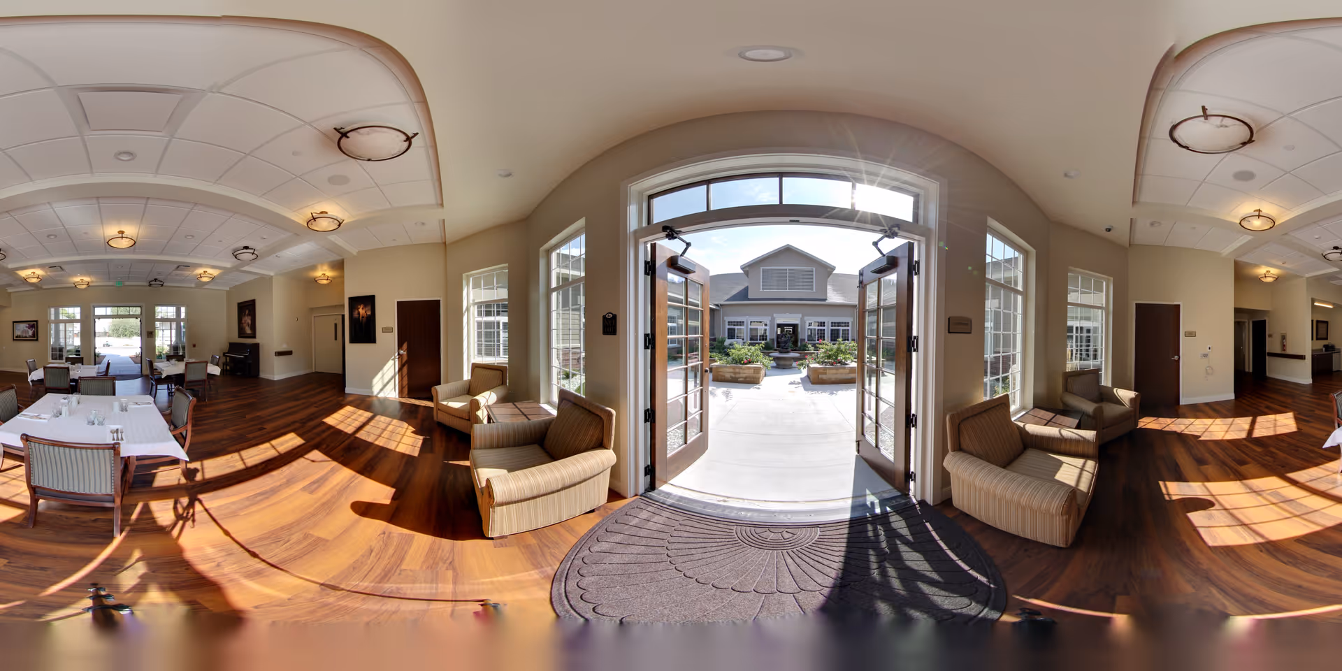Sunlit dining and lounge area with wooden floors, tables and chairs and armchairs by open double doors leading to an outdoor courtyard.