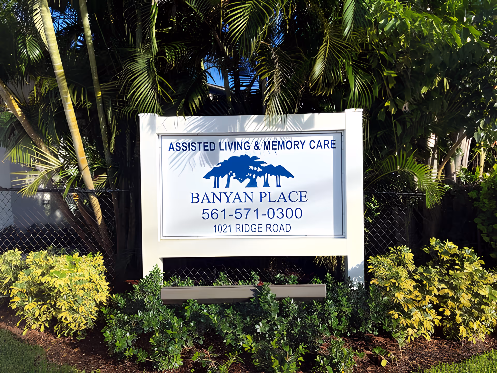A white signboard surrounded by green plants and palm trees. The sign reads 'Assisted Living & Memory Care, Banyan Place, 561-571-0300, 1021 Ridge Road' with a blue logo of a tree above the text.