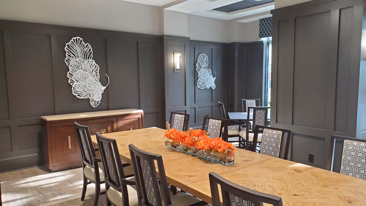 A dining room with a long wooden table surrounded by chairs, an orange floral centerpiece, dark paneled walls, and decorative wall art.