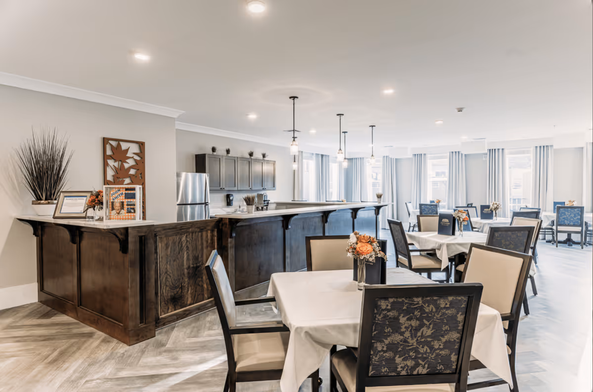 A bright and spacious dining room in a senior living facility with multiple tables covered in white tablecloths, each adorned with a small floral centerpiece. The room features large windows with light curtains, a wooden bar counter with pendant lights hanging above, and a stainless steel refrigerator in the background. The flooring is a light wood pattern, and the overall decor is modern and welcoming.