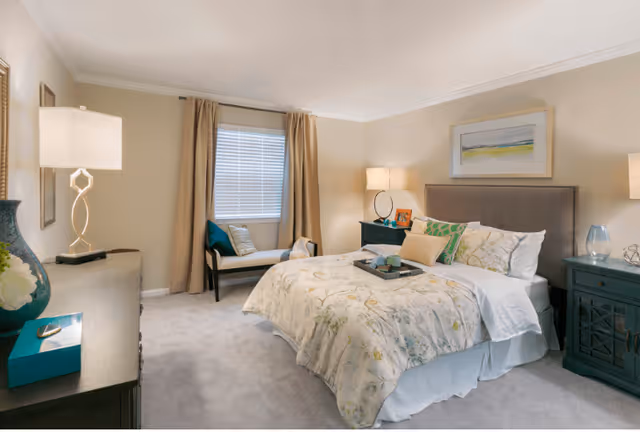 A cozy bedroom with a large bed covered in floral bedding and multiple pillows. There are two dark wooden nightstands on either side of the bed, each with a lamp. A window with beige curtains and a cushioned bench underneath is visible on the far wall. The room has light-colored walls and carpeted flooring, with framed artwork hanging above the bed.