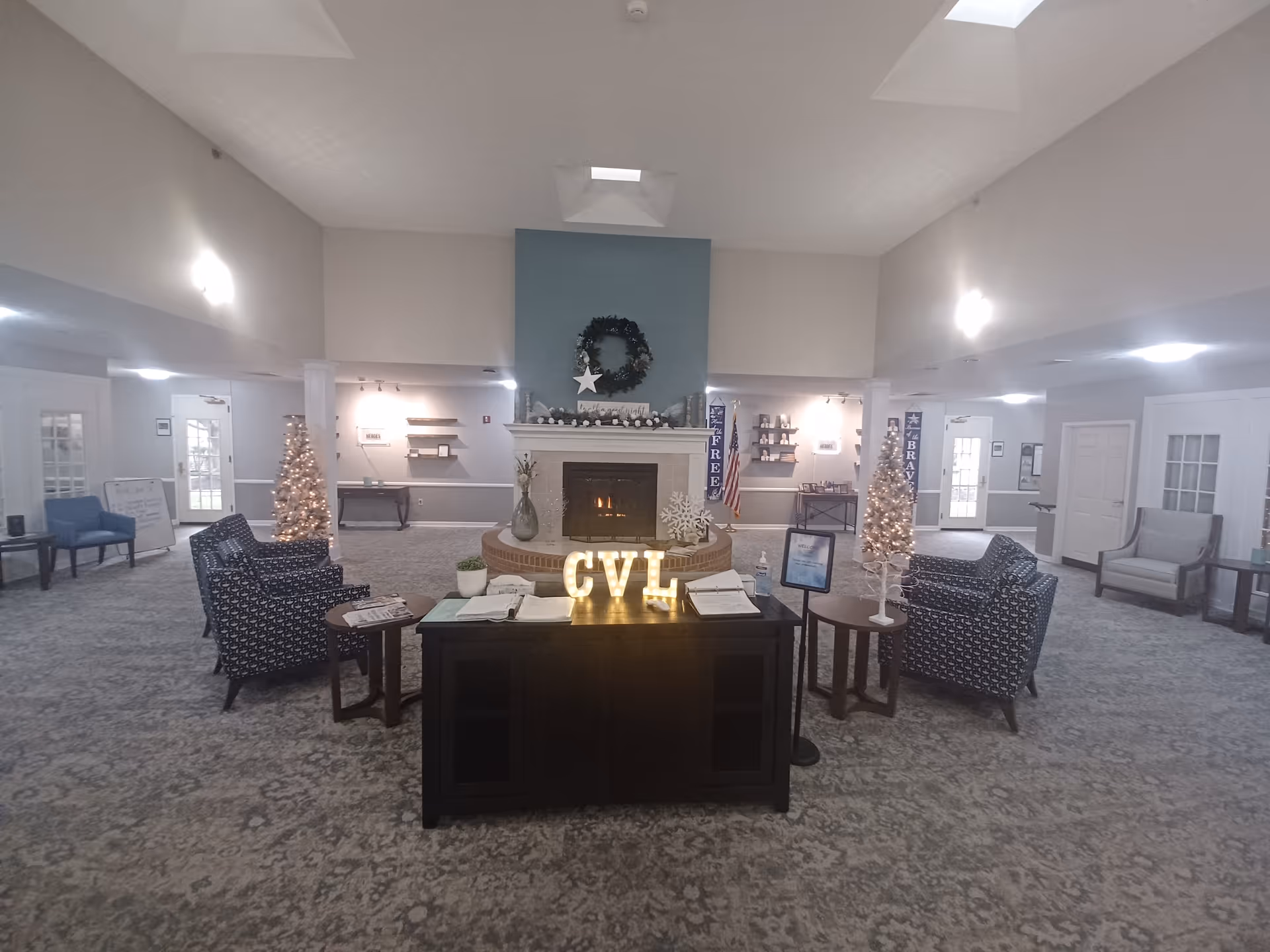 A spacious and well-lit common area with a fireplace in the center, decorated with a wreath and holiday ornaments. There are two decorated Christmas trees on either side of the fireplace. Comfortable armchairs and small tables are arranged around the room. A dark wooden cabinet with illuminated letters 'CVL' sits in front of the fireplace, along with open books and a hand sanitizer bottle. The room has a high ceiling with skylights and light gray walls.