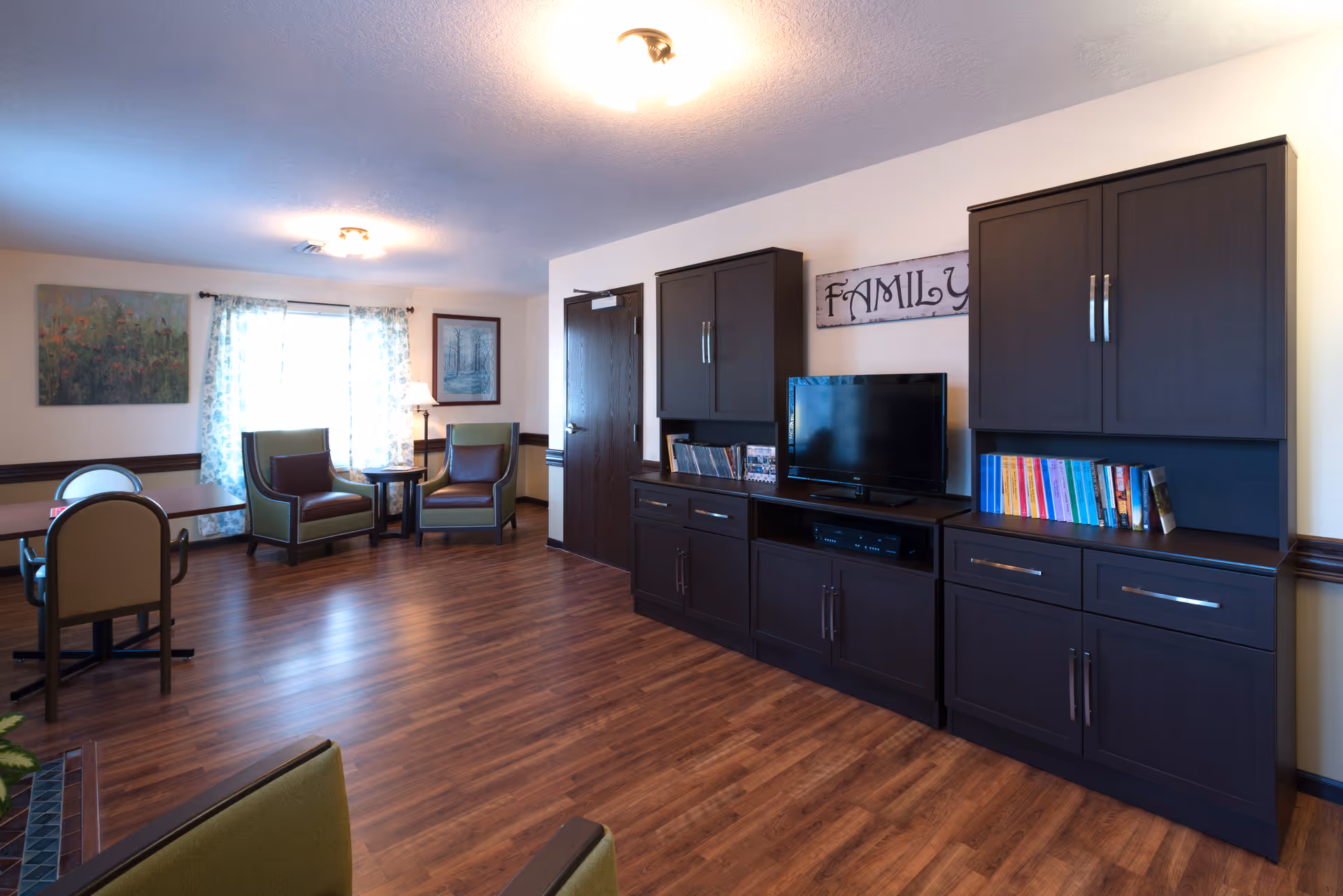 A spacious living room area with wooden flooring, two green armchairs with brown cushions, a small round table with a lamp between them, a large dark wood entertainment center with a TV, books, and cabinets, and a window with sheer curtains letting in natural light. The wall above the TV has a decorative sign that reads 'FAMILY'.