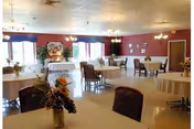 A spacious dining room with multiple round tables covered with white tablecloths and chairs around them. The walls are painted red with several windows letting in natural light. There are flower arrangements on some tables and a buffet table set up near the windows.