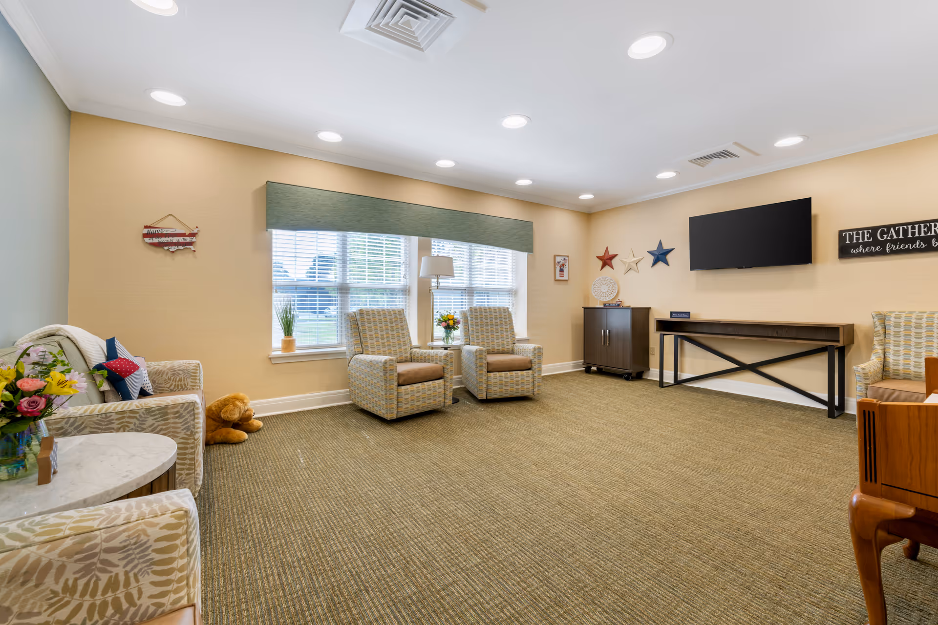 A bright and cozy living room in a senior living facility with beige walls and carpet. The room features several patterned armchairs arranged around the space, a flat-screen TV mounted on the wall, decorative stars and a sign that reads 'THE GATHERING where friends b...' on the wall. There are large windows with blinds and a green valance letting in natural light. A small cabinet and a console table are also present, along with a teddy bear on the floor and a vase of flowers on a side table.
