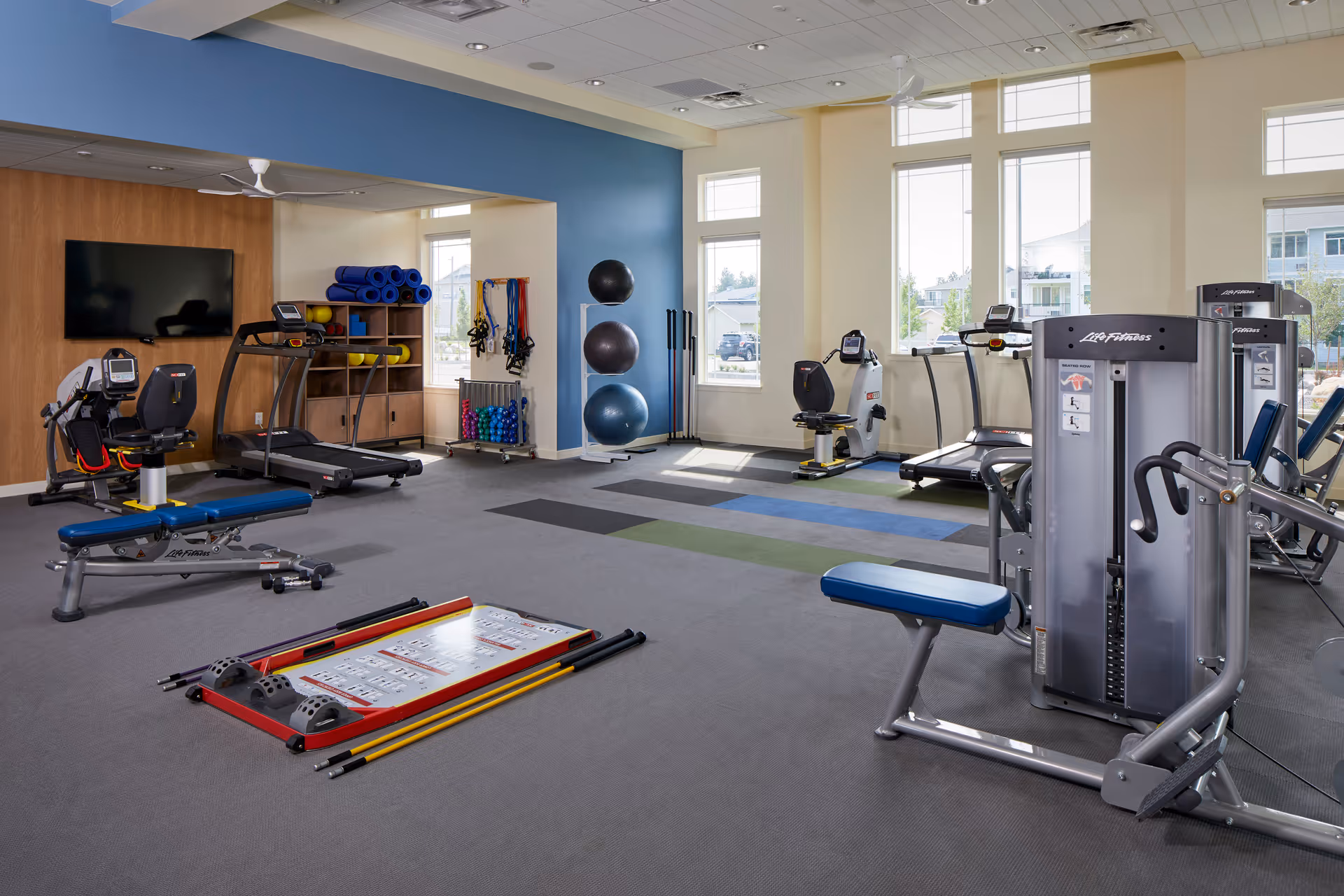 Well-lit fitness room with treadmills, weight machines, exercise balls and mats in front of large windows.