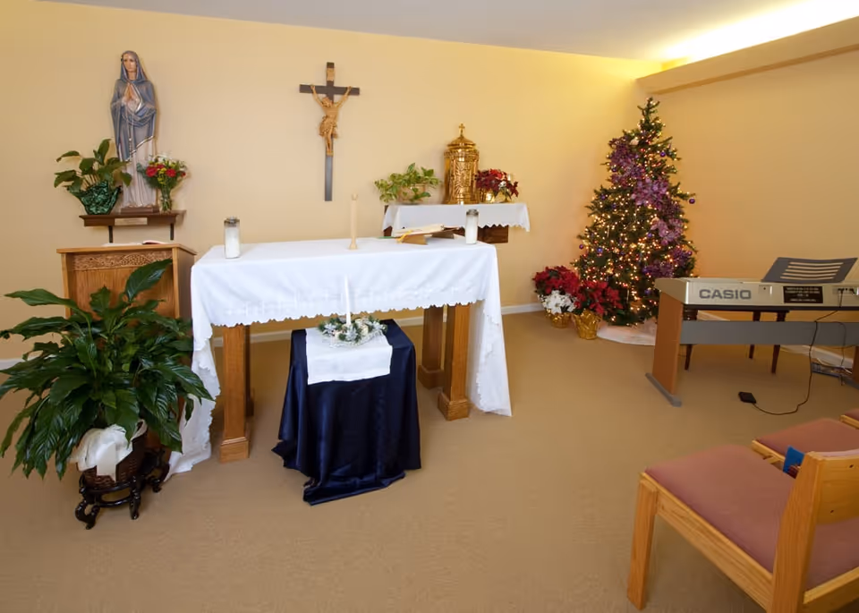 A small chapel room with a white altar covered with a cloth, candles, and religious items. A statue of the Virgin Mary and a crucifix are mounted on the wall. A decorated Christmas tree with lights and poinsettia plants are in the corner. A Casio keyboard and wooden chairs with cushions are also visible.