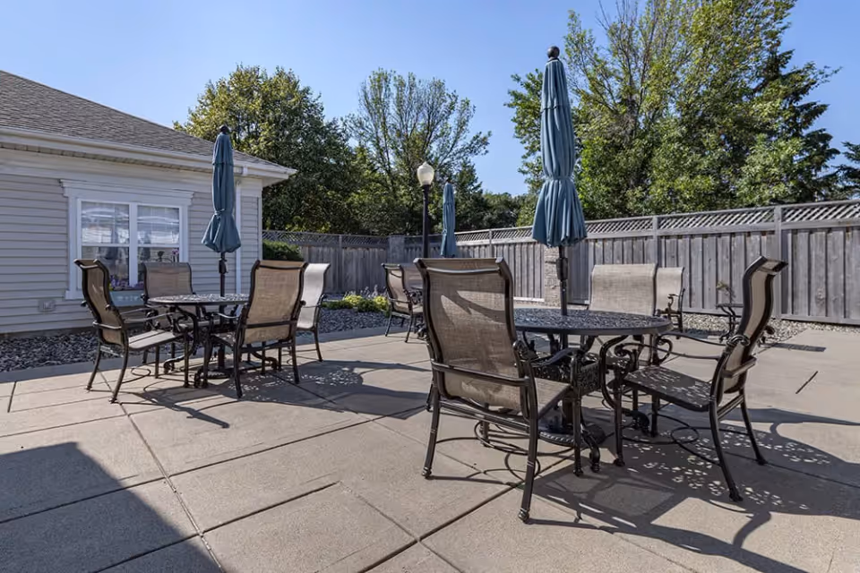 Outdoor patio area with multiple round metal tables and beige mesh chairs. Each table has a closed blue umbrella. The patio is surrounded by a wooden fence and there are trees and a building with windows in the background under a clear blue sky.