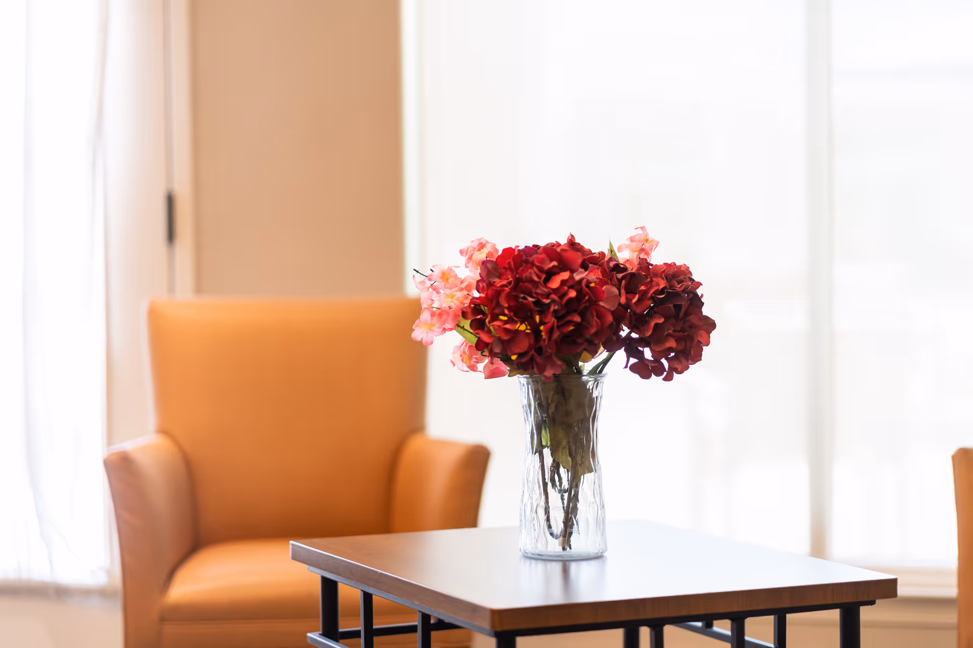 A clear glass vase with red and pink flowers sits on a wooden table in front of an orange armchair, with bright natural light coming through a window in the background.
