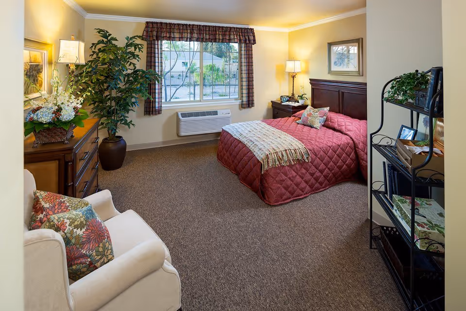 A cozy bedroom in a senior living facility featuring a bed with a red quilted bedspread and decorative pillows, a wooden nightstand with a lamp, a large window with plaid curtains, a potted plant, a wooden dresser with a floral arrangement, a comfortable armchair with floral cushions, and a black metal shelving unit with books and plants.