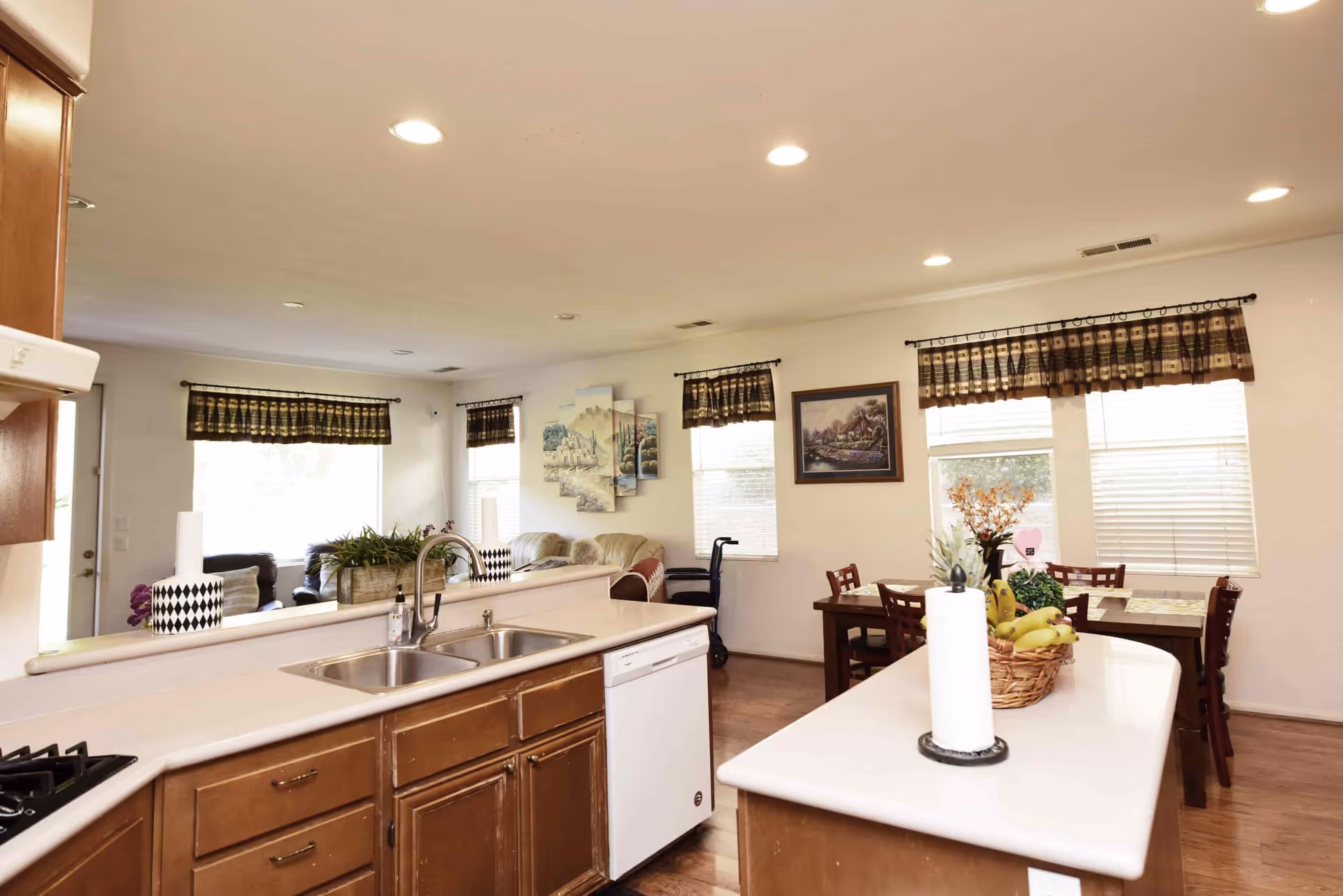 Bright and spacious kitchen area with wooden cabinets, a double sink, and a white dishwasher. The kitchen opens into a dining area with a wooden table and chairs, decorated with a basket of fruit and a paper towel holder on the island. The room has multiple windows with patterned valances, letting in natural light. In the background, there is a living area with sofas and wall art.