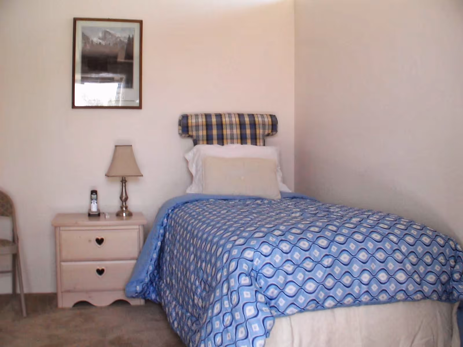 A small bedroom with a single bed covered in a blue patterned comforter, a nightstand with a lamp and framed artwork on the wall.