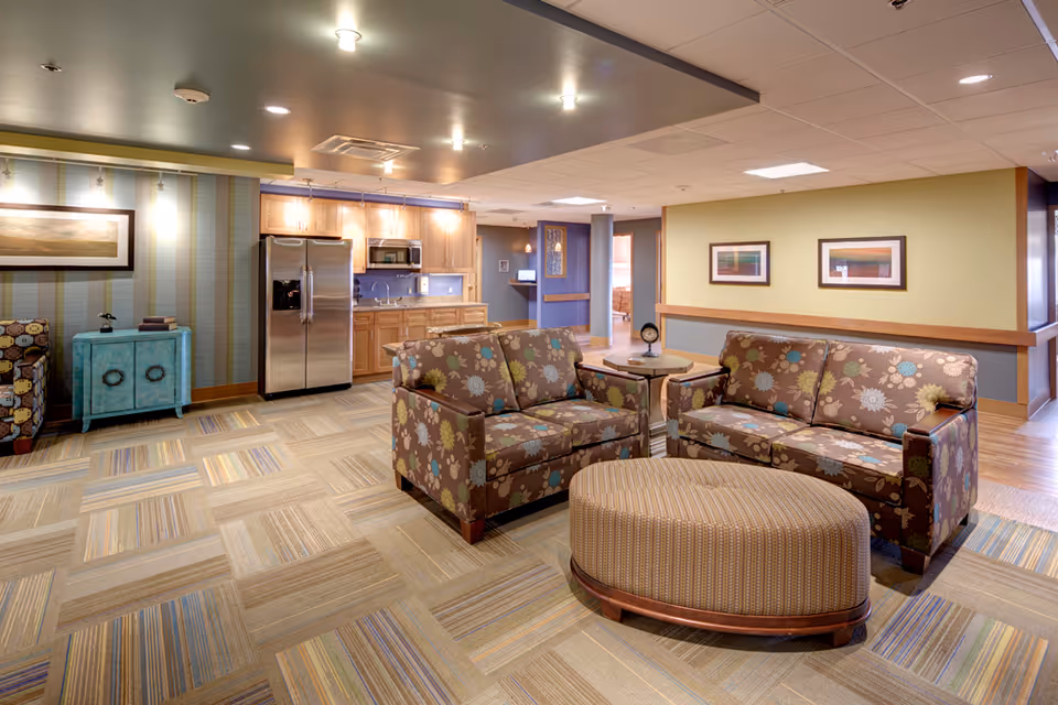 Bright common lounge with floral-patterned sofas, an ottoman, and a kitchenette with a stainless steel refrigerator.