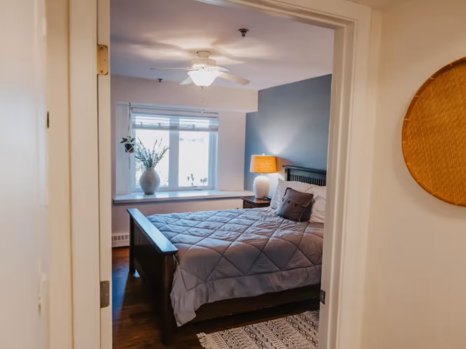 View through a doorway into a cozy bedroom with a bed, nightstand and lamp, window with a vase, and ceiling fan.