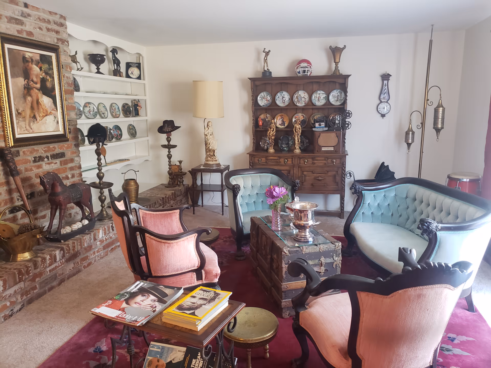A vintage-style living room with antique furniture including a light blue tufted sofa, matching armchair, and two pink upholstered chairs arranged around a wooden trunk coffee table. The room features a brick fireplace with decorative items, a wooden cabinet displaying decorative plates and figurines, a floor lamp, and a small table with magazines and books. The walls are white, and there is a red carpet on the floor.