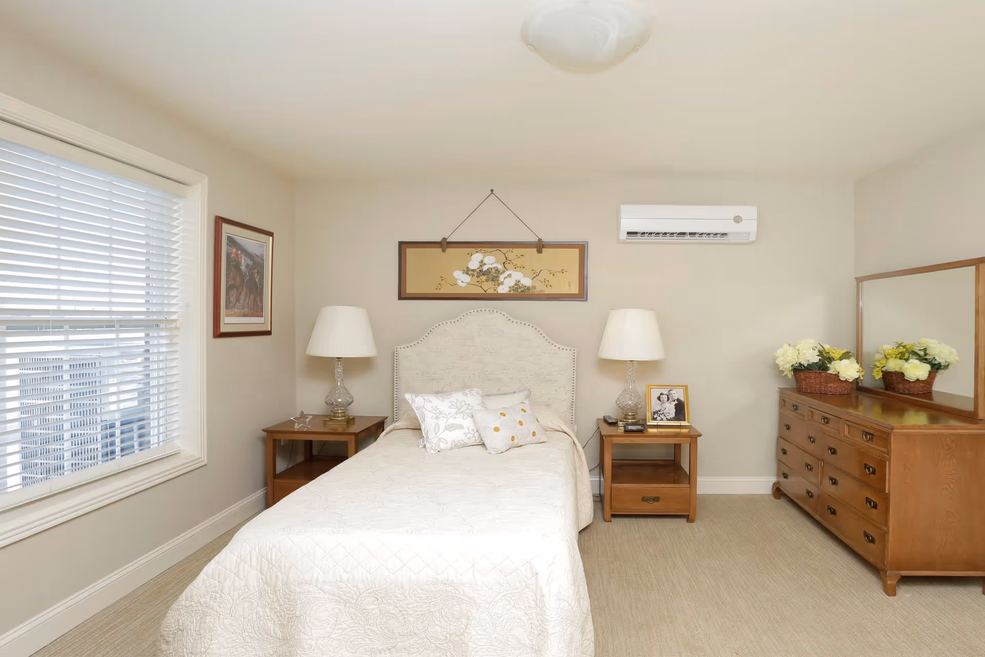 A neatly arranged bedroom with a single bed covered in a cream quilt and two decorative pillows. On either side of the bed are wooden nightstands with glass lamps. Above the bed hangs a framed floral artwork. To the right is a wooden dresser with a large mirror and two baskets of yellow flowers. A window with white blinds is on the left wall, and an air conditioning unit is mounted on the wall above the right nightstand.