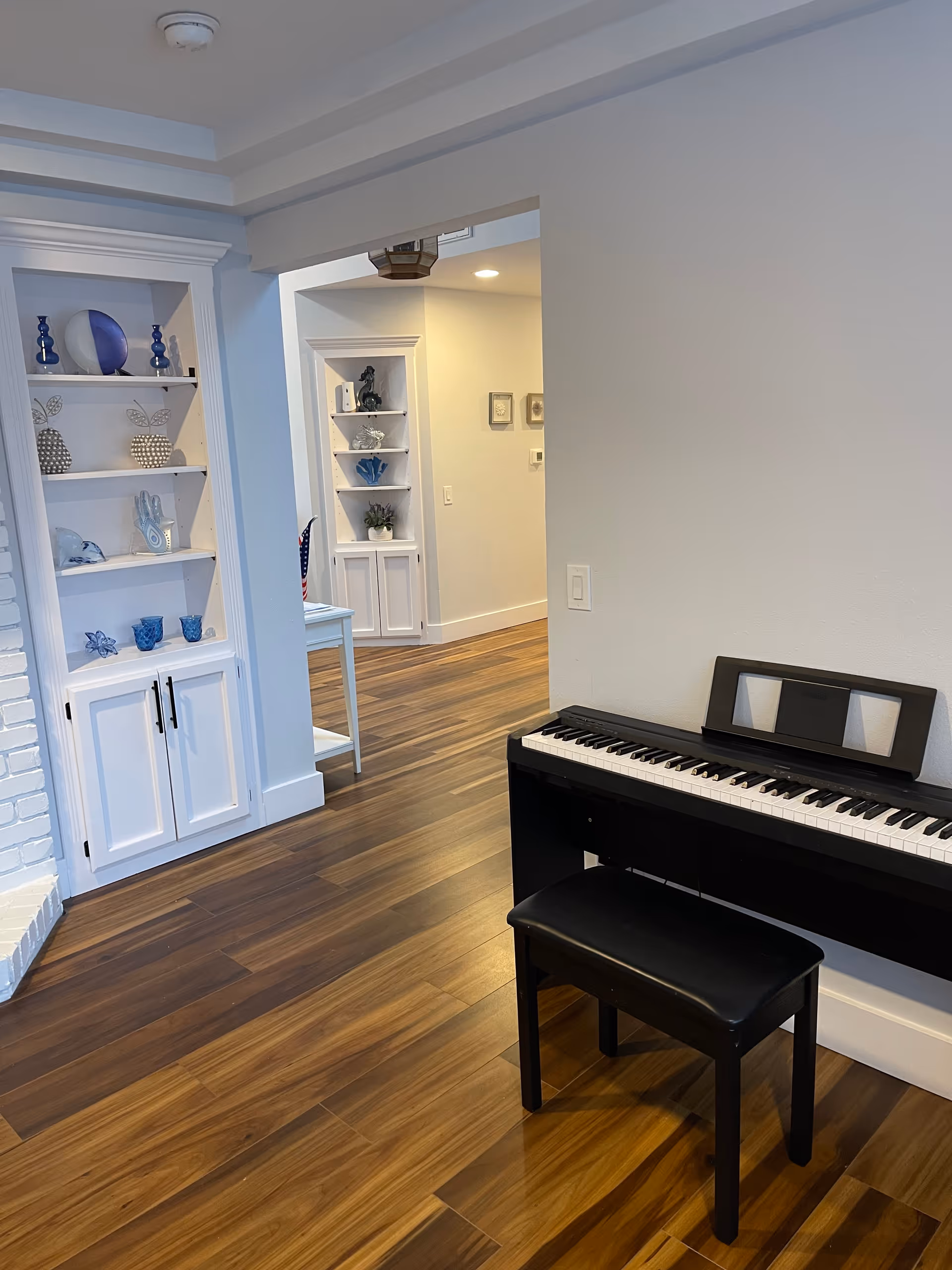 Interior view of a room with wooden flooring, a black electronic keyboard with a matching bench against a white wall, and built-in white shelves displaying decorative blue and white items. The room opens into a hallway with additional built-in shelves and light-colored walls.