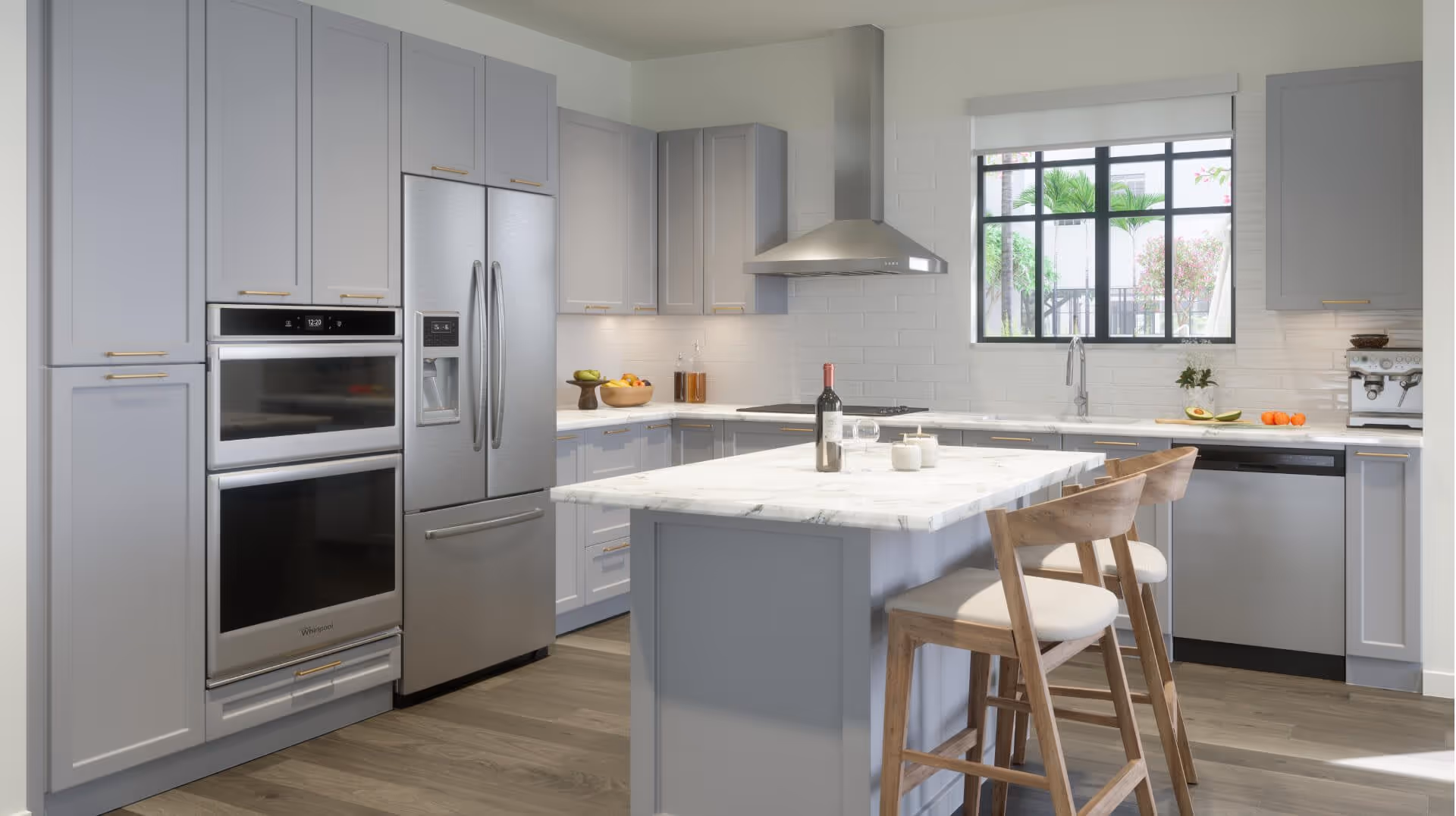 Modern kitchen with light gray cabinets, stainless steel double oven and refrigerator, a kitchen island with marble countertop, two wooden bar stools with white cushions, a window with black frame showing greenery outside, and various kitchen items including a bottle of wine, glasses, fruit bowls, and a coffee machine.