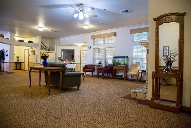 A spacious living room area with patterned carpet, a ceiling fan with lights, and multiple seating options including a green sofa, red and beige chairs. There is a wooden table with a blue vase of yellow flowers, a fish tank on a wooden stand, and a large wooden mirror with decorative carvings. The room has large windows with blinds and valances, and soft lighting from ceiling fixtures.