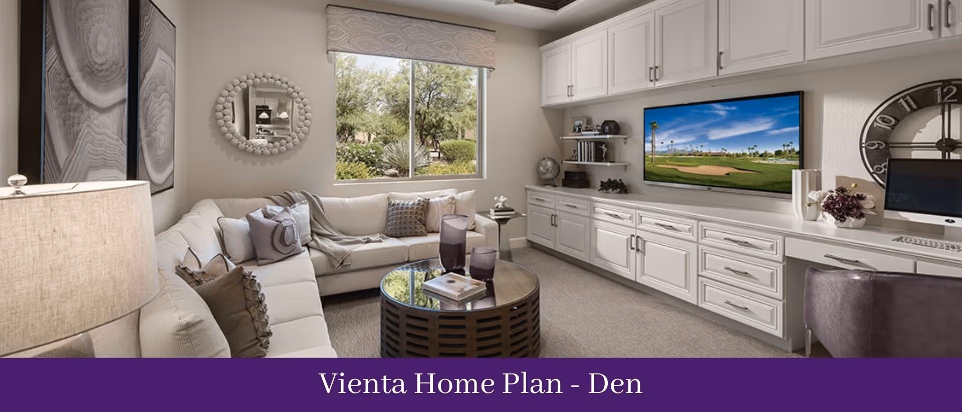A cozy den featuring a large beige sectional sofa with multiple cushions, a round coffee table with decorative items, a window showing greenery outside, built-in white cabinetry with a mounted flat-screen TV displaying a golf course, and a desk area with a computer and a large wall clock.