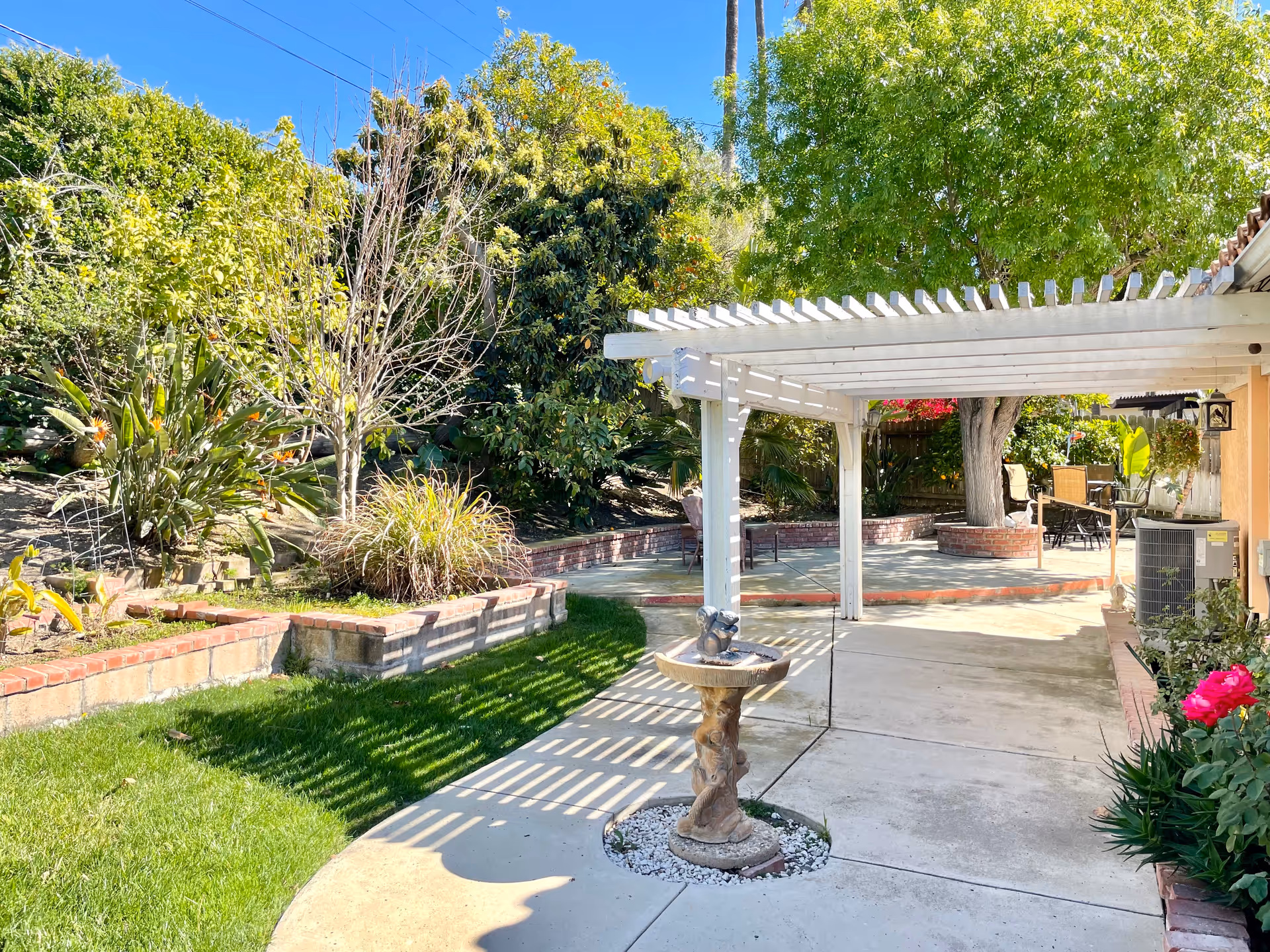 Outdoor garden area with a white pergola casting shadows on a concrete pathway. There is a small decorative birdbath in the foreground surrounded by white stones. The garden has green grass, various plants, trees, and brick-bordered flower beds. Patio furniture is visible in the background under the shade of trees.