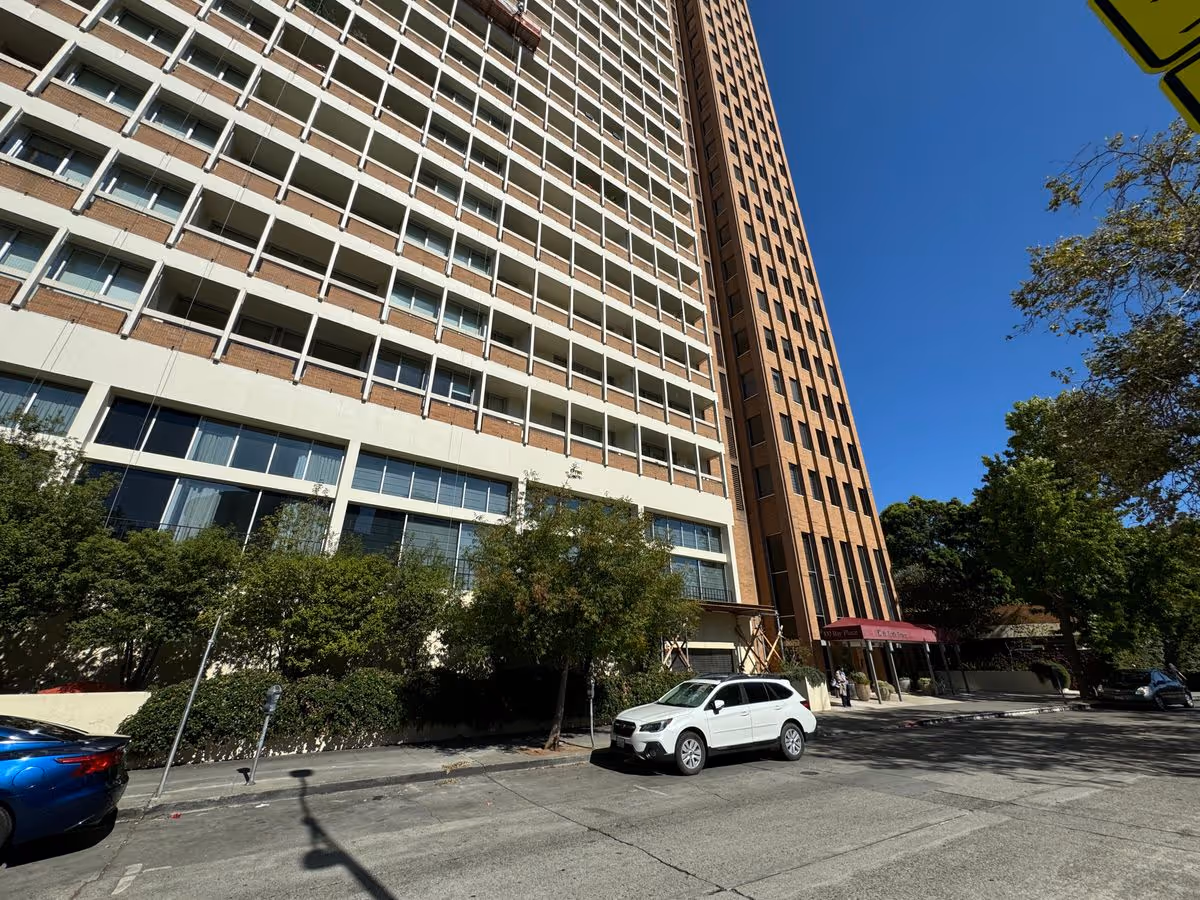 Street-level view of a tall multi-story residential building with balconies, trees, and parked cars under a clear blue sky.