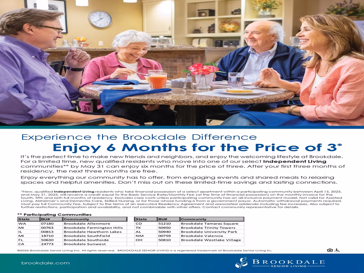 Four seniors sitting around a round dining table in a well-lit room, smiling and enjoying a meal together. The table has plates, glasses, a small flower vase, and blue napkins. The background shows a kitchen area with cabinets and a clock on the wall.