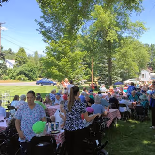 A large group of people gathered outdoors under trees, sitting at tables covered with red and white checkered tablecloths, enjoying a social event on a sunny day.