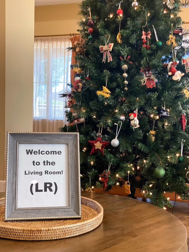 A decorated Christmas tree beside a framed sign reading "Welcome to the Living Room! (LR)" on a table in a living room.