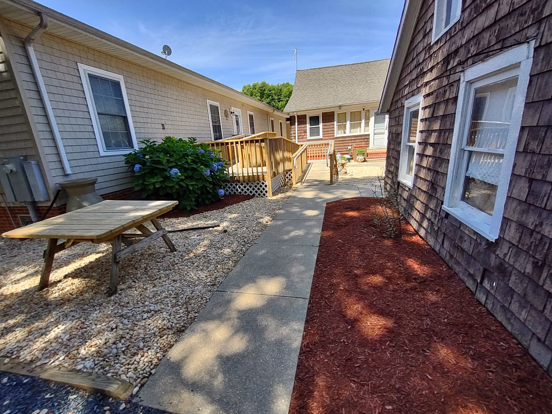 Outdoor courtyard area between buildings at Whispering Pines Assisted Living Facility with a concrete walkway, a wooden picnic table on gravel, a wooden ramp, and landscaped areas with mulch and plants under a clear blue sky.
