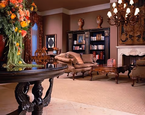 Elegant living room with classic furniture including a carved wooden table with a large floral arrangement, a vintage sofa and armchair, a wooden coffee table, a bookshelf filled with books and decorative items, a fireplace with a mantel, and a chandelier hanging from the ceiling.