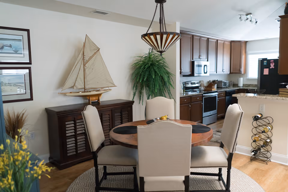 A dining area with a round wooden table surrounded by four beige upholstered chairs. Behind the table is a dark wooden cabinet with a decorative sailboat model on top and two framed pictures on the wall. A large green fern plant hangs on the wall near the kitchen, which features wooden cabinets, stainless steel appliances, and a granite countertop. A wine rack with several bottles is placed near the kitchen counter.