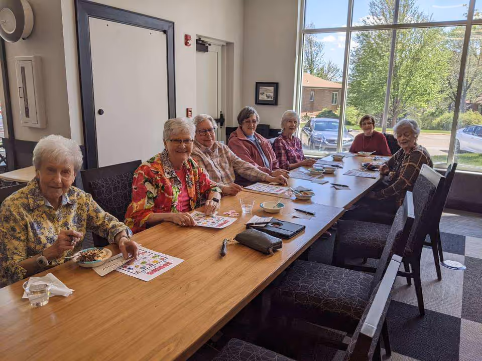 Seven elderly women seated around a long table in a bright community dining/activity room with large windows.
