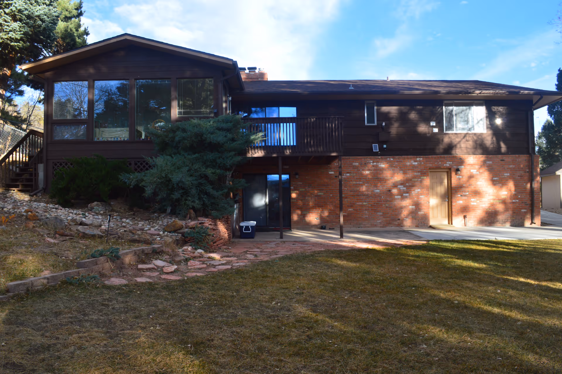 Exterior view of a two-story residential building with a brick lower level and dark wood upper level. The building features large windows, a small balcony, a door on the lower level, and a grassy yard with some landscaping and trees around it under a blue sky.