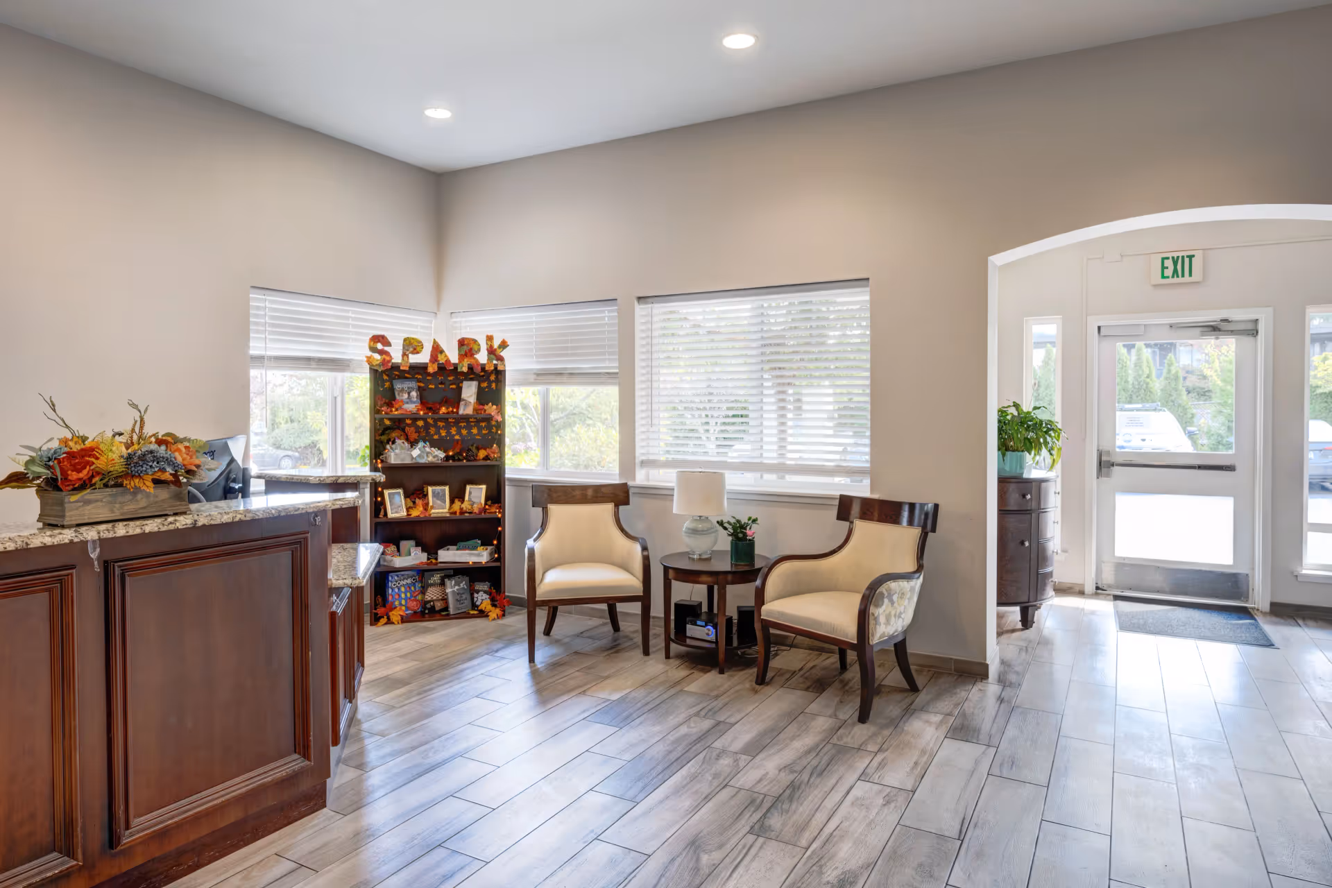 Bright reception lobby with a wooden front desk, two upholstered chairs around a small table, decorative shelving, and an exit door.