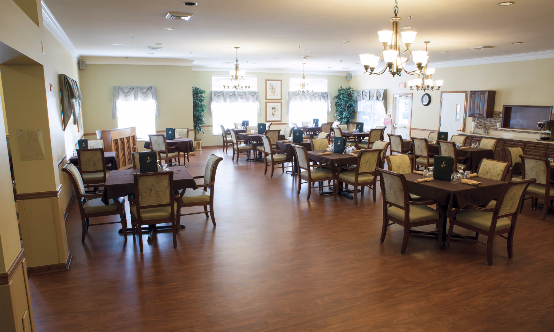 A spacious dining room with multiple tables covered in dark tablecloths, each surrounded by wooden chairs with cushioned seats and backs. The room has large windows with valances, two chandeliers hanging from the ceiling, and plants placed near the windows. There is a piano against one wall and a counter area with cabinets and a coffee machine on the right side.