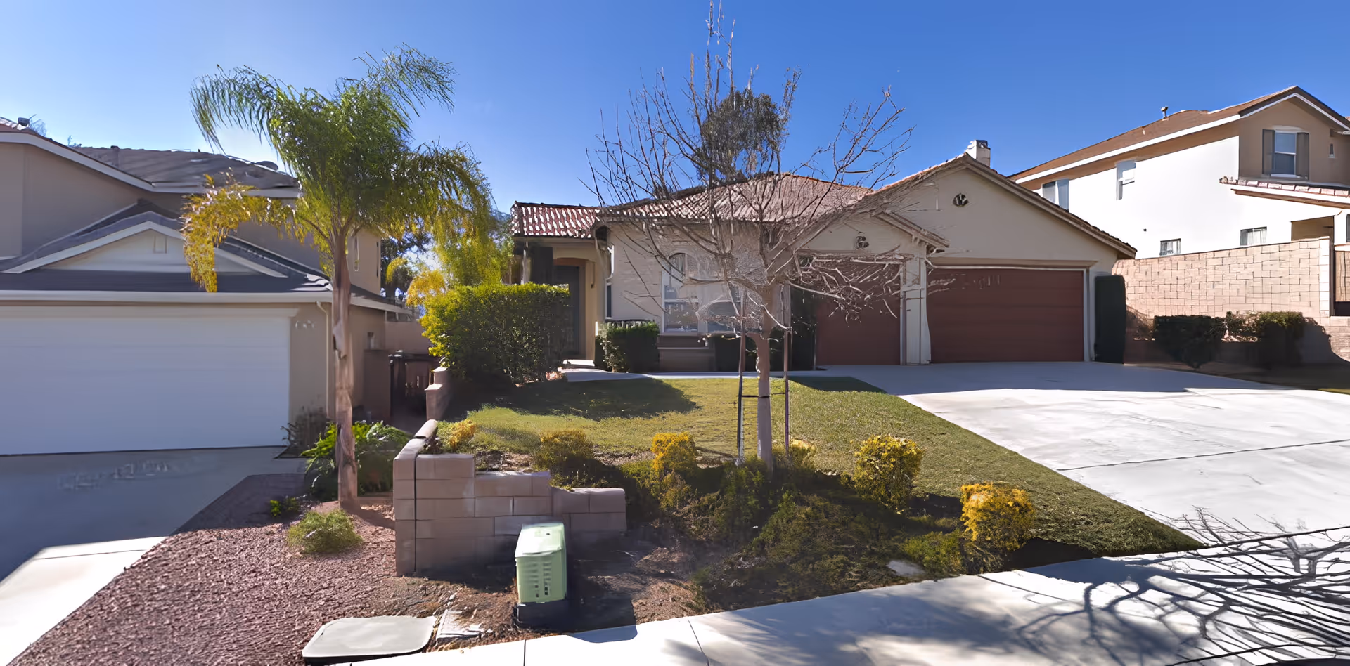 Front exterior view of a single-story residential building with a two-car garage, a driveway, a small lawn, and some landscaping including a palm tree and bushes under a clear blue sky.
