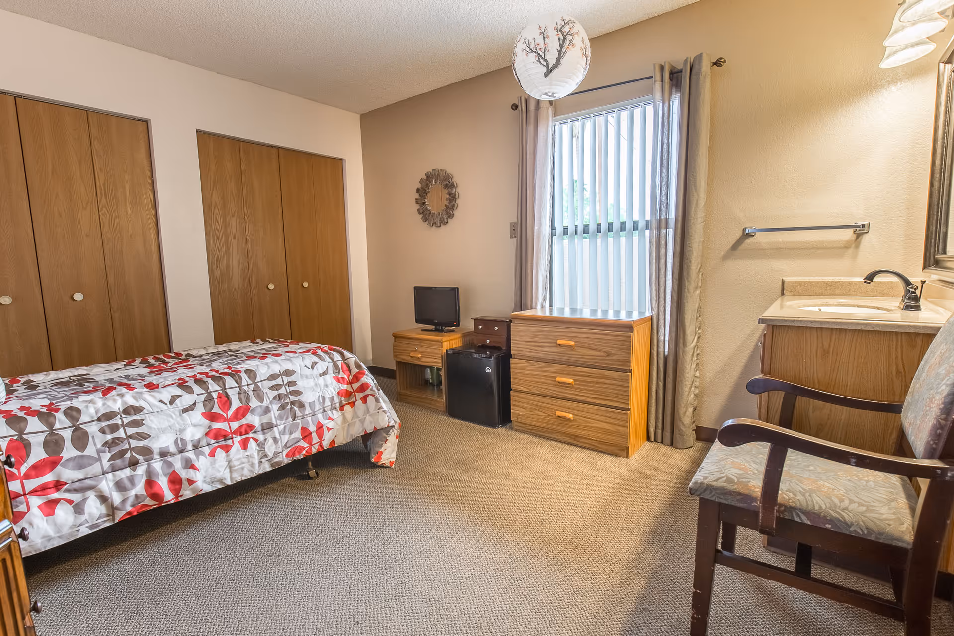 A bedroom in an assisted living facility featuring a single bed with a floral patterned bedspread, a wooden dresser, a small TV on a stand, a mini refrigerator, a chair with floral upholstery, a sink with a mirror above it, and a window with vertical blinds and curtains.