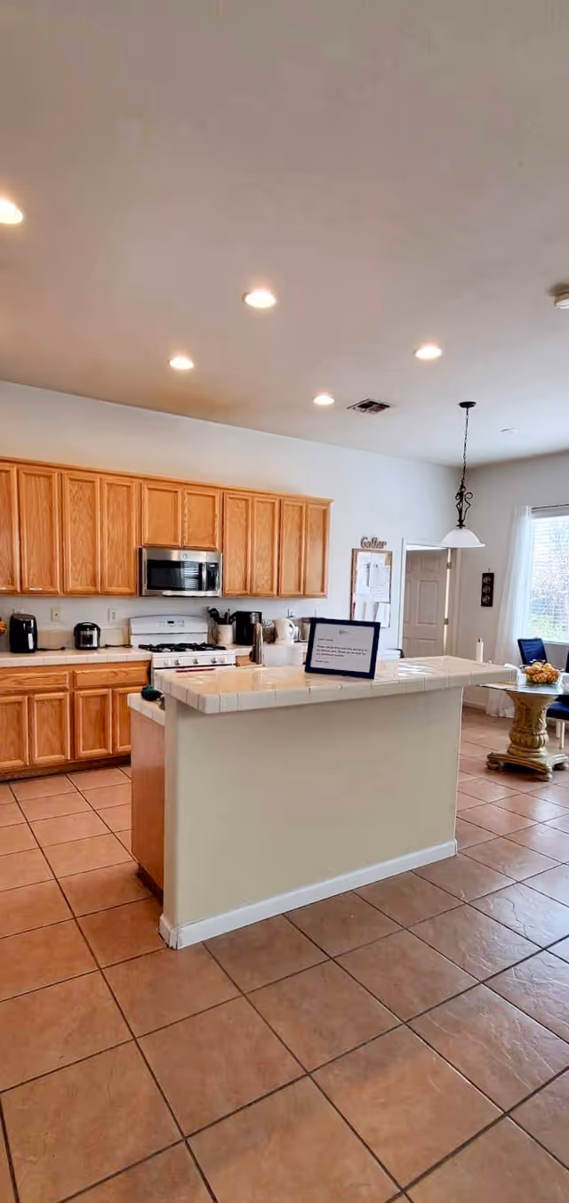 A bright kitchen with wooden cabinets, a white tiled countertop island, and stainless steel microwave above a stove. The floor is tiled with large brown tiles. There are several kitchen appliances on the counter, and a hanging light fixture is visible near a window with white curtains. A small table with chairs is seen in the background near the window.